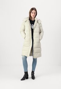 Light beige puffer coat with a hood, long length, button closure, quilted texture, worn over a black graphic shirt and blue jeans. Black ankle boots.