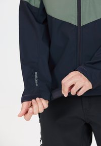 Marineblå og grøn vandtæt jakke med lynlås foran, justerbare manchetter og velcro-lukninger. Har "W-PRO 9000" branding på ærmet.