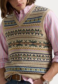 Stickad väst i beige med flerfärgade geometriska mönster, med V-ringning och ribbad fåll; lagd över en ljusrosa randig skjorta.