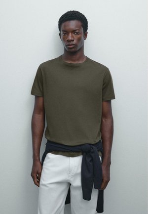 Olijfgroene katoenen T-shirt met een ronde hals, korte mouwen; gelaagd met een marineblauwe trui gestrikt rond de taille over witte broek.