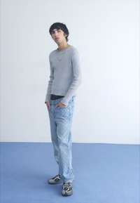 Lys grå tettsittende genser, svart undertrøye, lyseblå slitte jeans og flerfargede joggesko. Glatt tekstur, avslappet passform, synlige sømmer.