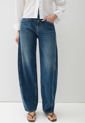 Vrouw draagt losse blauwe jeans, wit overhemd met knopen en groene teenslippers, staand met één hand in haar zak tegen een effen achtergrond.