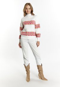 Maglione bianco a maglia intrecciata con motivi geometrici rosa e gialli, abbinato a jeans beige e stivali da cowboy color beige. Texture liscia, collo alto.