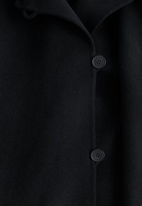 Juodas vilnonis paltas su įpjovu apykakle ir dviem juodais mygtukais. Audinys yra lygaus paviršiaus ir turi minimalų dekoravimą.