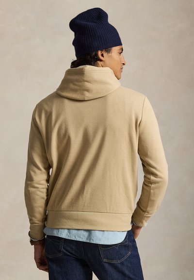 Bărbat purtând un hanorac bej, cămașă bleu deschis, blugi închiși la culoare și o căciulă tricotată bleumarin, orientat lateral, cu un fundal neutru.