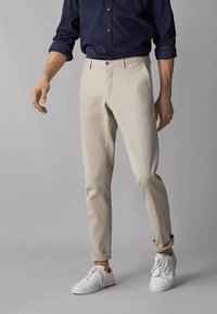 Beige bomullschinos med slim fit, med sidofickor och en enkel knappstängning, parat med vita sneakers mot en neutral bakgrund.