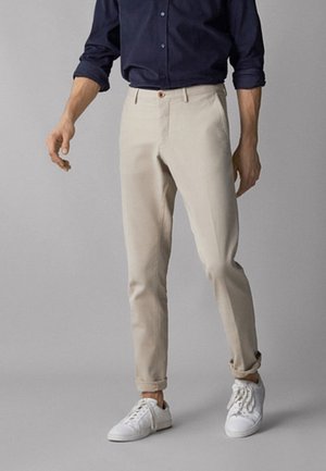 Chinos in cotone beige con una vestibilità slim, dotati di tasche laterali e chiusura a un solo bottone, abbinati a sneaker bianche su uno sfondo neutro.