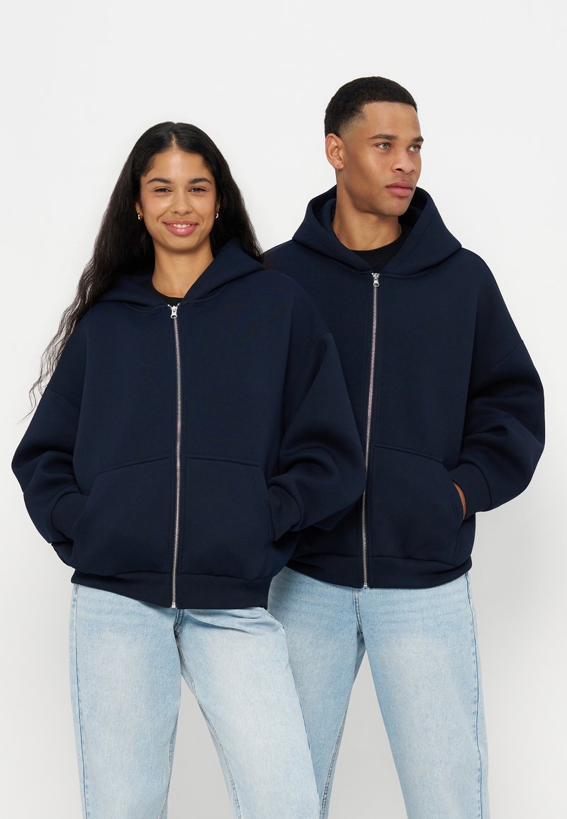Deux adultes portant des sweats à capuche bleu marine zippés et des jeans bleu clair sont côte à côte sur un fond blanc.