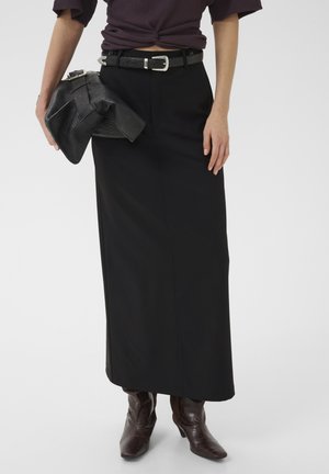 Person iført et sort højtaljet langt skørt, brune støvler med hæl og holder en sort tekstureret clutch taske.