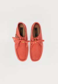 Mocasines de ante color coral con bordes cosidos, detalle de lazo y acentos de cuentas de madera; diseñados para la comodidad y el uso casual.