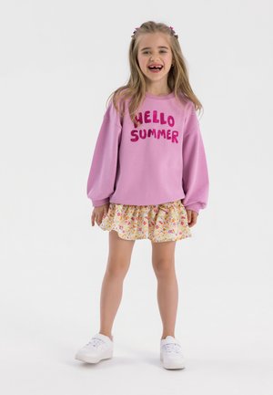 Rosa sweatshirt med "HELLO SUMMER" i glittrande bokstäver, matchad med en blommönstrad kjol med volanger. Vita sneakers fulländar looken.