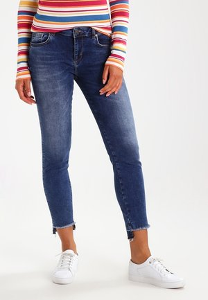 Person iført mørkeblå skinny jeans med frynsete kanter, en farverig stribet langærmet top og hvide snørebåndssneakers.