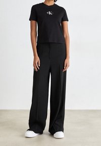 Sort cropped t-shirt med hvid CK-logo, parret med løse sorte bukser med sidelommer og hvide sneakers. Glatte teksturer hele vejen igennem.