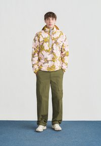 Νεαρός άνδρας που φοράει ροζ και κίτρινο φλις μπουφάν με σχέδια, χακί παντελόνι cargo και λευκά αθλητικά παπούτσια, όρθιος πάνω σε μπλε χαλί.