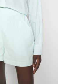 Haut bleu clair à manches longues assorti à un short bleu clair, taille élastique, main reposant à côté de la cuisse sur fond uni.