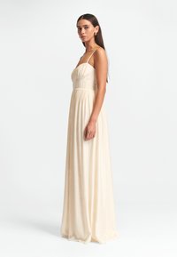 Vestido maxi beige con tirantes finos, cuerpo ajustado y tela fluida; presenta una sutil textura y frunces en la cintura.