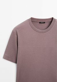 T-shirt a maniche corte e collo rotondo in cotone di un tenue colore malva. Presenta una texture liscia con una sottile cucitura orizzontale sul petto.