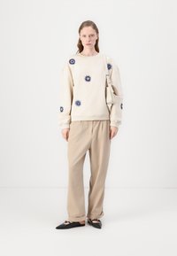 Beige sweatshirt med blå solmotiv, tillsammans med lösa beige byxor och svarta platta skor, kompletterat med en liten vit väska.