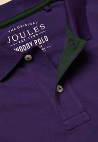Violeta polo krekls ar zaļu apkakli, kurai ir pogu aizdare un teksturēts audums. Logotips un zīmola nosaukums ir redzams apkakles iekšpusē.