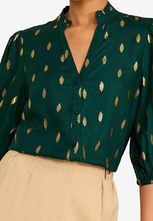 Blouse vert foncé avec motif à pois dorés, boutonnée sur le devant, manches trois-quarts bouffantes, associée à un pantalon beige taille haute.