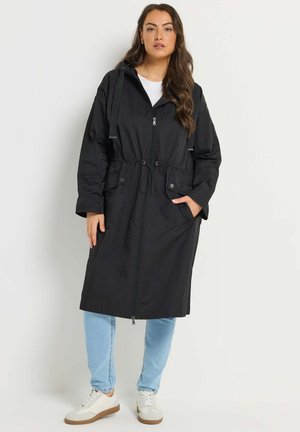 Vrouw die een lange zwarte capuchonjas draagt met een ritssluiting aan de voorkant, een aantrekkoord in de taille, zakken, lichtblauwe spijkerbroek en witte sneakers, staand tegen een witte muur.