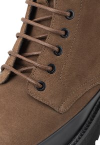 Bota de tobillo de ante marrón con ojales negros y cordones marrones, que presenta una parte superior texturizada y una suela de goma negra. Vista en primer plano de los detalles.