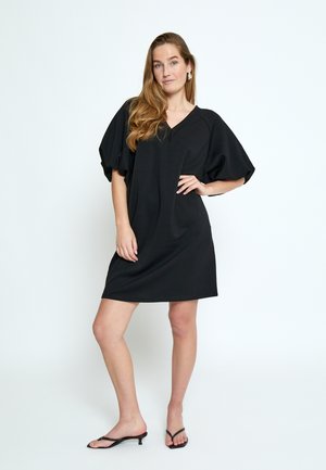 Mujer con el pelo largo, vestida con un vestido negro de escote en V, mangas abullonadas y tacones negros abiertos en la punta, de pie con una mano en la cadera sobre un fondo blanco.