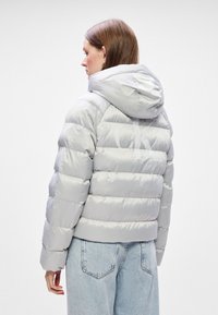 Svijetlosivi puffer s kapuljačom, s kviltanim prugama i logom na leđima, u kombinaciji sa svijetloplavim trapericama.