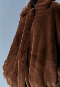 Hnědá bunda z umělé kožešiny s měkkou texturou, límcem a volným střihem, s kulatými rukávy a bez viditelných zapínání.