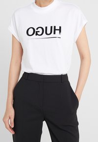 Camiseta blanca de algodón con mangas enrolladas, con el texto "HUGO" en negro y una línea horizontal. Combinada con pantalones negros a medida.