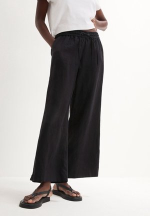Persona che indossa pantaloni neri ampi e larghi con coulisse in vita, t-shirt corta bianca e sandali neri con platform su sfondo bianco.