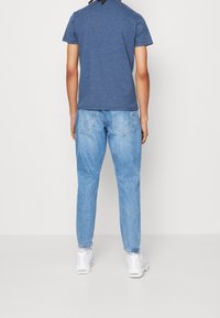 Persona vestida con jeans de mezclilla azul claro, camisa azul de manga corta y zapatillas blancas, de pie frente a una pared blanca lisa.