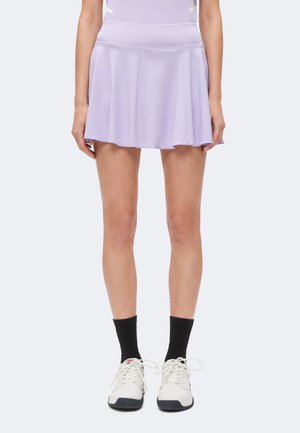 Personne portant une jupe évasée violet clair, des chaussettes noires montantes et des baskets blanches, debout devant un fond uni.