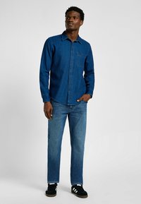 Niebieska dżinsowa koszula zapinana na guziki z kieszonką na piersi, w połączeniu z niebieskimi dżinsami i czarnymi trampkami. Gładka tekstura i standardowy krój kołnierza.