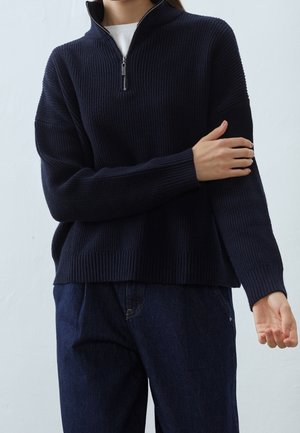 Personne portant un pull en maille côtelée bleu marine avec un col à demi-fermeture éclair et un jean bleu foncé sur un fond uni.