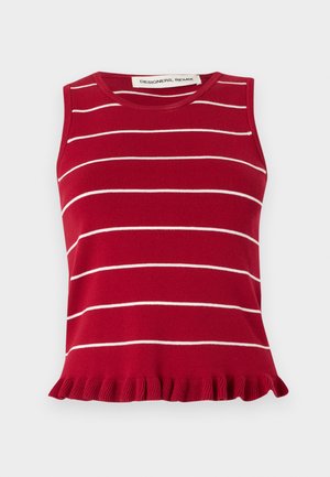 Γυναίκα στέκεται με σταυρωμένα χέρια, φοράει αμάνικο κόκκινο φόρεμα με λευκές ρίγες και βολάν στο τελείωμα, συνδυασμένο με μαύρες σαγιονάρες.