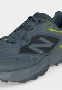 Σκούρο γκρι αθλητικό παπούτσι New Balance με επάνω μέρος από δίχτυ, εμφανές λογότυπο "N", κορδόνια και υφασμένο πέλμα με νέον πράσινες λεπτομέρειες.