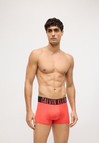 Crvene bokserice od rastezljivog materijala s crnim pojasom na kojem se ističe logo "CALVIN KLEIN" velikim slovima. Klasični prilagođeni dizajn.