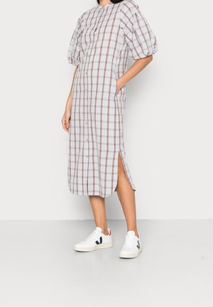 Kvinde iført hvid midi-kjole med brun ternet mønster, puffede ærmer, sideslids og hvide sneakers med sorte og røde detaljer.