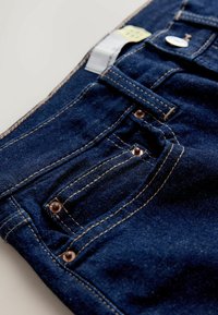 Jeans din denim de un albastru închis, cu un buzunar frontal prevăzut cu nituri de cupru și o cusătură contrastantă de culoare bej. Materialul are o textură netedă și o ușoară strălucire.