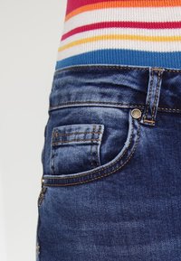 Jeans i mörkblå denim med en blekt textur, med en sidoficka och kontrasterande gyllene sömmar. Förstärkt med en metallknapp.