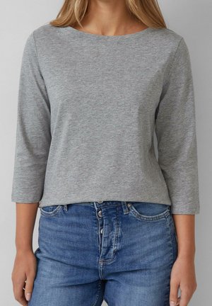 Femme portant une chemise à manches longues gris clair avec des manches trois-quarts et un jean bleu taille haute avec fermeture à boutons.