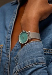 Montre avec un bracelet en maille argentée et un cadran rond vert menthe, présentant des lignes verticales et des index horaires argentés sur un fond en denim.