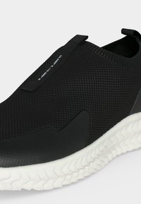 Baskets noires slip-on avec empeigne en mesh texturé, languettes de tirage noires et semelle blanche à motif ondulé visible de profil.
