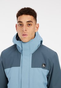 Giacca da sci azzurro chiaro con accenti blu scuro, colletto alto, chiusura con zip e logo sulla parte anteriore. Tessuto liscio, materiale resistente all'acqua.