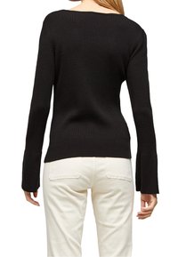 Svart ribbad stickad tröja med bred U-ringning och trumpetärmar, lagd över ljusa byxor; detaljerad textur synlig.