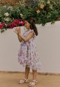Lichtpaarse jurk met bloemen- en vlinderpatronen, gerimpelde bandjes en een gelaagde rok. G gedragen met witte sandalen met een strikdetail.