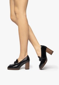 Mocasines de charol negro con tacón de bloque de madera texturizada, que presentan un lazo decorativo en la punta y un acabado brillante y suave.