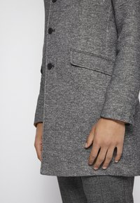 Manteau en laine gris avec une finition texturée, six boutons noirs et une poche latérale. Porté sur un pantalon à carreaux, présentant une coupe allongée.