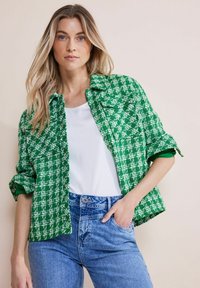 Grön och vit mönstrad jacka med krage, med knäppningar och uppvikta ärmar, bärs över en vit topp och blå jeans.